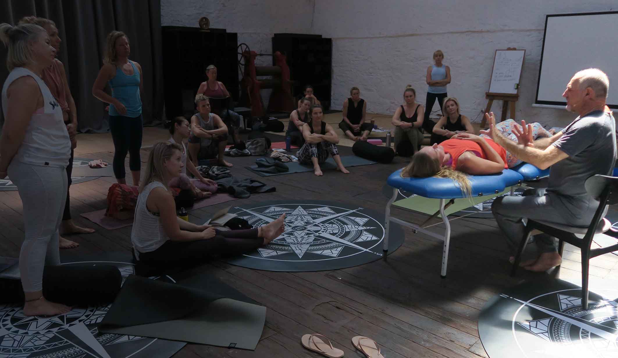 Leslie seated at a massage table demonstrating a technique for a roomful of students